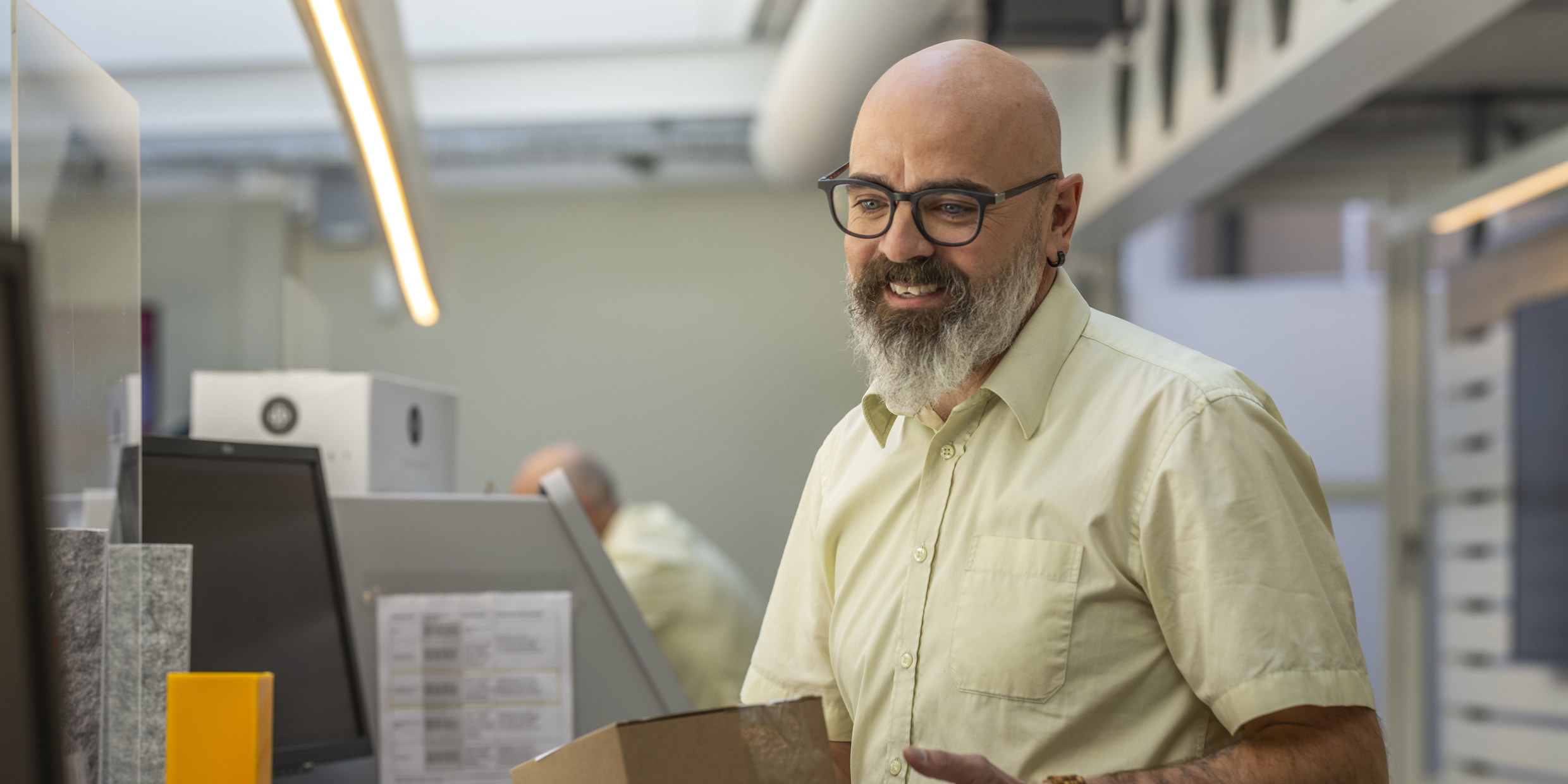 Ein Postbeamter mit Glatze, Brille und gelbem Hemd arbeitet am Schalter und hat ein Paket in der Hand